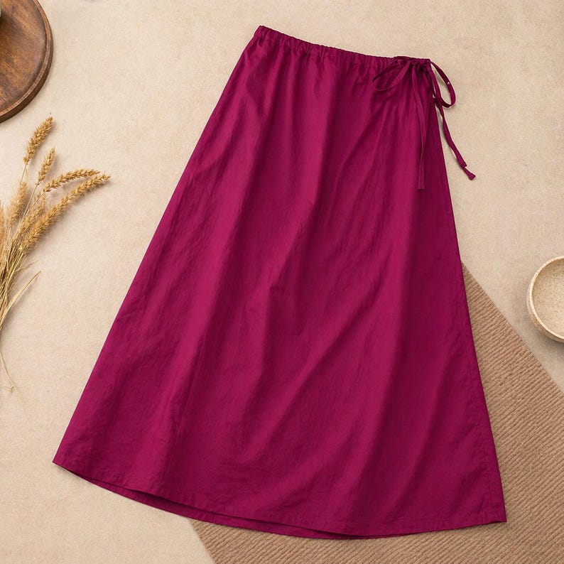 May include: A long, magenta skirt with a drawstring waist and tie closure. The skirt is made of a lightweight fabric and has a flowing silhouette. The skirt is a solid color and is suitable for casual wear.