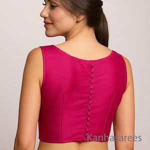May include: A magenta sleeveless blouse with a row of small buttons down the back. The blouse has a scoop neckline and is made of a smooth fabric. The text "Kanhasarees" is visible on the lower right.