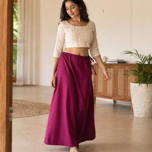 May include: A woman wearing a cream-colored, long-sleeved crop top and a long, magenta skirt. The skirt is tied at the waist with a matching magenta drawstring. The outfit is simple and elegant, suitable for casual or semi-formal occasions.