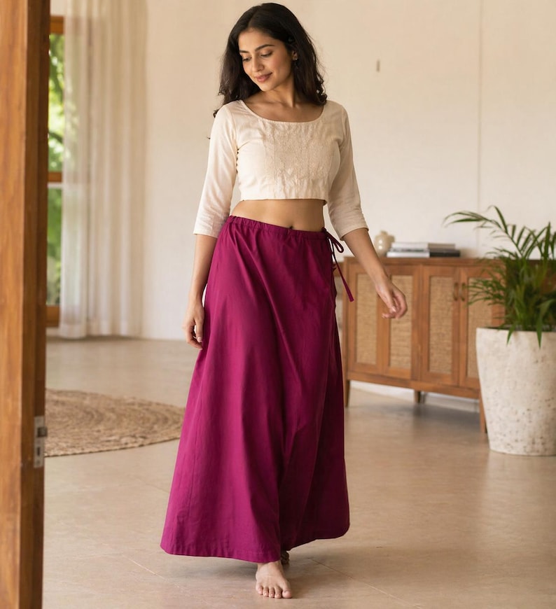 May include: A woman wearing a cream-colored, long-sleeved crop top and a long, magenta skirt. The skirt is tied at the waist with a matching magenta drawstring. The outfit is simple and elegant, suitable for casual or semi-formal occasions.
