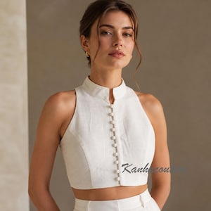 Peut inclure: Haut court sans manches blanc cassé avec un col montant et une rangée de petits boutons sur le devant. Le haut est en tissu texturé et a une coupe ajustée. Le texte "Kanhcou" est visible en bas à droite.