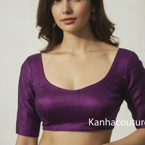May include: A dark purple blouse with a scoop neckline and three-quarter sleeves. The blouse has a subtle sheen and is paired with a light purple skirt. The text "Kanhacouture" is visible on the lower right.