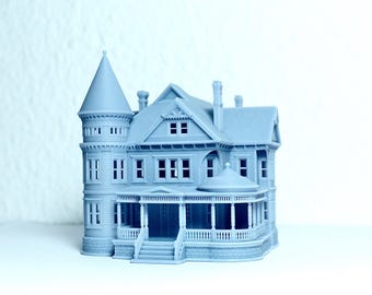 May include: A light blue miniature Victorian house model with a turret, porch, and multiple windows. The house has a detailed design, including a front porch with columns and a staircase leading to the entrance. The model is a detailed architectural replica.