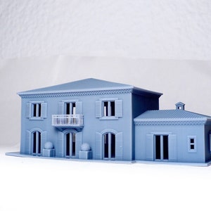 Pu&ograve; includere: Un modello di casa a due piani di colore blu chiaro stampato in 3D con un balcone e una struttura pi&ugrave; piccola annessa. La casa ha modanature e finestre bianche.