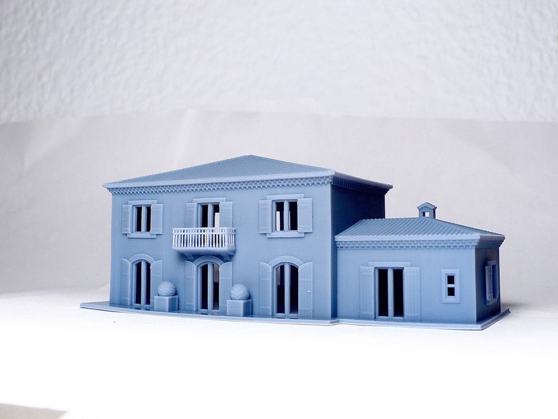 Pu&ograve; includere: Un modello di casa a due piani di colore blu chiaro stampato in 3D con un balcone e una struttura pi&ugrave; piccola annessa. La casa ha modanature e finestre bianche.