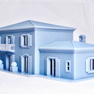 Pu&ograve; includere: Un modello di casa piccola stampato in 3D di colore azzurro con un balcone bianco, persiane e un camino. La casa ha un design semplice con una forma rettangolare e un tetto a due falde.