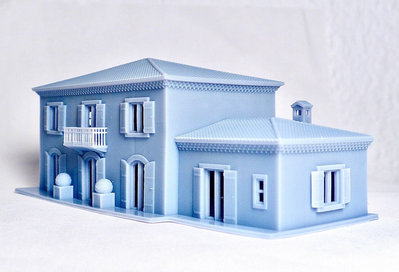Pu&ograve; includere: Un modello di casa piccola stampato in 3D di colore azzurro con un balcone bianco, persiane e un camino. La casa ha un design semplice con una forma rettangolare e un tetto a due falde.
