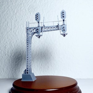 Può includere: Un modello grigio stampato in 3D di un ponte di segnalazione ferroviaria caratterizzato da un'alta torre di supporto a traliccio con una scala attaccata e un braccio orizzontale a sbalzo. Include quattro testate di segnale.