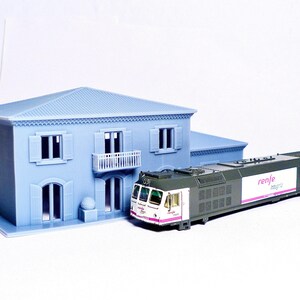Pu&ograve; includere: Un set di treni in miniatura con un treno bianco e verde con la scritta "renfe" sul lato. Il treno &egrave; parcheggiato davanti a una casa in miniatura blu con finiture bianche.