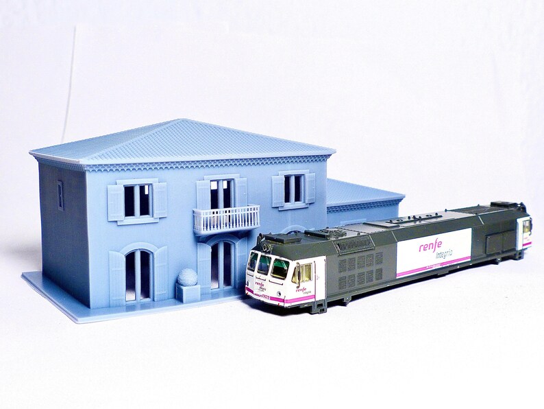 Pu&ograve; includere: Un set di treni in miniatura con un treno bianco e verde con la scritta "renfe" sul lato. Il treno &egrave; parcheggiato davanti a una casa in miniatura blu con finiture bianche.
