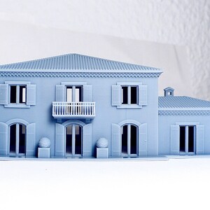 Pu&ograve; includere: Un modello di casa a due piani stampato in 3D di colore grigio con un balcone, persiane e un'ala separata. Il modello &egrave; posizionato su una superficie bianca.
