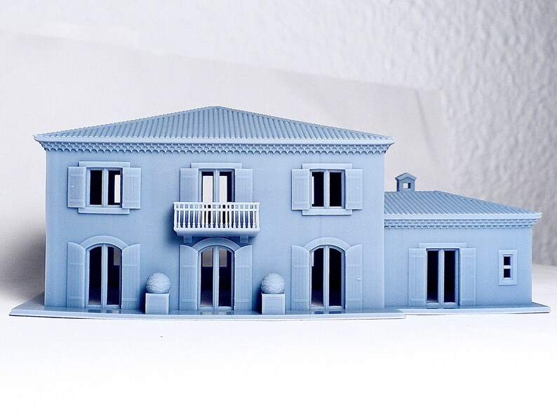 Pu&ograve; includere: Un modello di casa a due piani stampato in 3D di colore grigio con un balcone, persiane e un'ala separata. Il modello &egrave; posizionato su una superficie bianca.