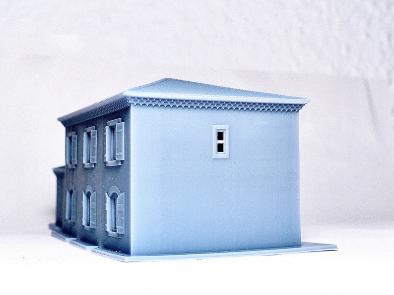 Pu&ograve; includere: Un modello 3D stampato in grigio chiaro di una piccola casa con un design semplice. La casa ha un tetto piatto, alcune finestre e una piccola porta. Il modello &egrave; realizzato in un materiale liscio e opaco.