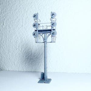 May include: A gray model of a stadium light tower. The tower has a rectangular base, a cylindrical pole, and a top section with multiple circular lights. The lights are arranged in two rows, with three lights in each row.