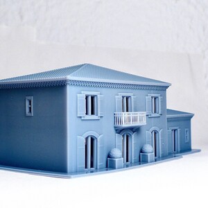 Pu&ograve; includere: Un modello di casa a due piani stampato in 3D di colore azzurro con un balcone, finestre e una porta. La casa ha una ringhiera bianca sul balcone e una cornice bianca attorno alle finestre e alla porta.