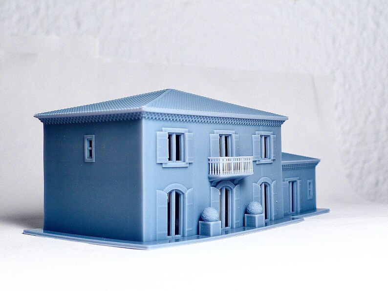 Pu&ograve; includere: Un modello di casa a due piani stampato in 3D di colore azzurro con un balcone, finestre e una porta. La casa ha una ringhiera bianca sul balcone e una cornice bianca attorno alle finestre e alla porta.