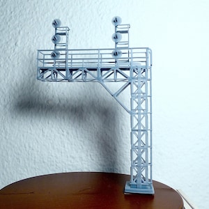 Może przedstawiać: Szary model wieży sygnalizacji kolejowej z wieloma światłami. Konstrukcja ma prostokątną podstawę, pionowe podparcie i poziomą platformę z barierkami. Światła są małe, okrągłe i równomiernie rozmieszczone.