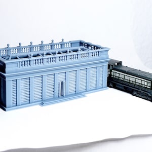 May include: A gray model of a building with a balcony and a green model train next to it.
