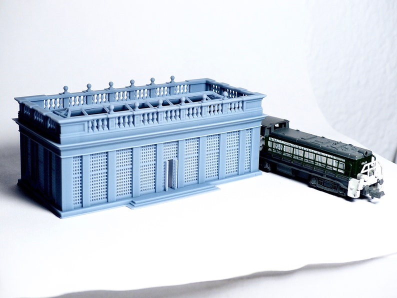 May include: A gray model of a building with a balcony and a green model train next to it.