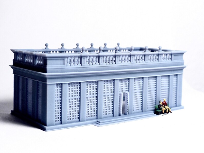 May include: A gray 3D printed model of a building with a balcony and a grid pattern on the windows. Two small figures are sitting on a bench in front of the building.