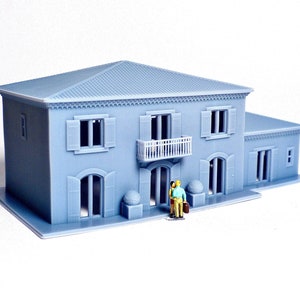 Pu&ograve; includere: Un modello di casa a due piani di colore grigio stampato in 3D con un balcone, finestre e una porta. Due piccole figure si trovano davanti alla casa.