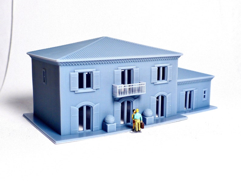 Pu&ograve; includere: Un modello di casa a due piani di colore grigio stampato in 3D con un balcone, finestre e una porta. Due piccole figure si trovano davanti alla casa.