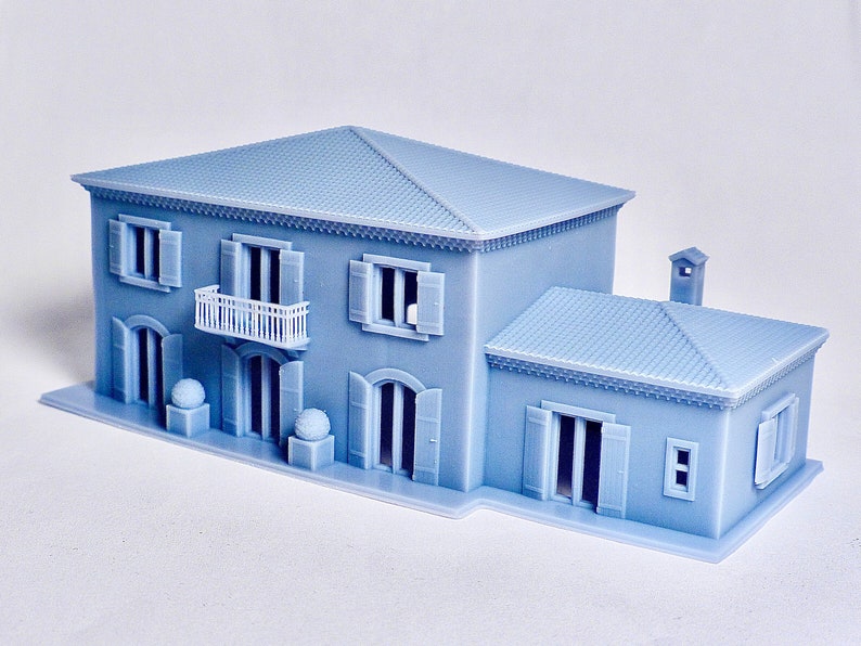 Pu&ograve; includere: Un modello di casa a due piani stampato in 3D di colore azzurro con un balcone bianco, un piccolo camino e un tetto in tegole.