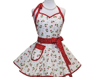 May include: A white apron with a red cherry pattern, red trim, and a red polka dot waist tie. The apron has a halter neck and a pocket. The apron is displayed on a mannequin.