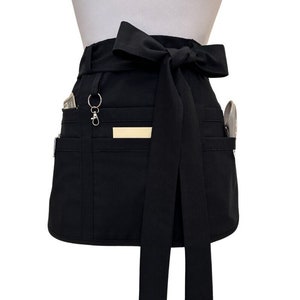 May include: Black waist apron with multiple pockets and a long tie. The apron has a silver-colored key ring and a small rectangular tag. A few items are visible in the pockets, including a utensil and some cash.
