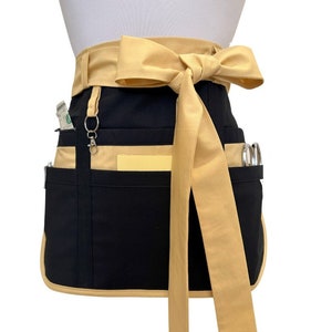 May include: A black and tan waist apron with multiple pockets and a large bow. The apron has a tan waistband and trim, with a black body and pockets. A silver-colored clip and a few items are visible in the pockets.