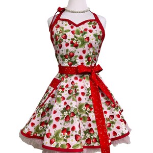 May include: A pink apron with a strawberry pattern, red trim, and a matching red bow. The apron has a halter neck and a full skirt, with a white tulle hem. The design features red strawberries, green leaves, and white flowers.