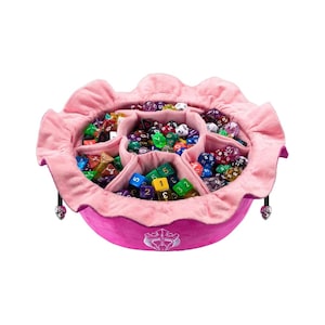 K&ouml;nnte beinhalten: Ein rundes W&uuml;rfeltablett aus rosa Stoff mit ger&uuml;schtem Rand und internen Trennw&auml;nden, gef&uuml;llt mit einer gro&szlig;en Sammlung bunter polyedrischer W&uuml;rfel. Die Au&szlig;enseite zeigt ein wei&szlig;es gesticktes Emblem und zwei kleine silberne Totenkopf-Anh&auml;nger.