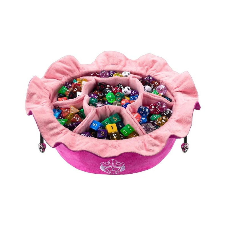 K&ouml;nnte beinhalten: Ein rundes W&uuml;rfeltablett aus rosa Stoff mit ger&uuml;schtem Rand und internen Trennw&auml;nden, gef&uuml;llt mit einer gro&szlig;en Sammlung bunter polyedrischer W&uuml;rfel. Die Au&szlig;enseite zeigt ein wei&szlig;es gesticktes Emblem und zwei kleine silberne Totenkopf-Anh&auml;nger.