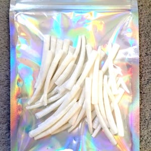 May include: A collection of numerous small, curved, white cylindrical pieces contained within a clear, iridescent, holographic plastic resealable pouch. The pouch features a single circular hole at the top for hanging and displays a rainbow sheen.