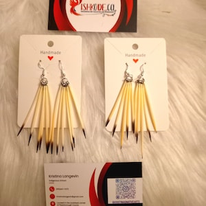 May include: Handmade earrings with a natural, earthy aesthetic. Each earring features multiple light-colored, slender elements with dark tips, suspended from silver-toned hooks. The image also includes business cards with the text "ISHKODE.CO" and "Kristina Langevin Indigenous Artisan."
