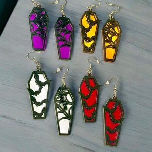 May include: Six pairs of coffin-shaped earrings with a bat design. The earrings are available in red, white, purple, and yellow.
