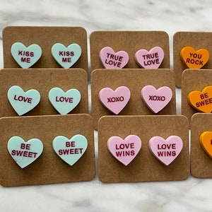May include: Assortment of heart-shaped earrings with various sayings, including "Kiss Me", "True Love", "You Rock", "Love Wins", "Be Sweet", and "Hug Me". The earrings are displayed on brown card stock.