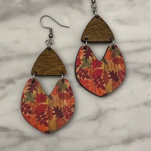 May include: A pair of wooden earrings with a fall leaf design. The earrings are made of two pieces of wood, one with a triangle shape and the other with a heart shape. The heart-shaped piece is decorated with a print of fall leaves in shades of red, orange, and yellow.