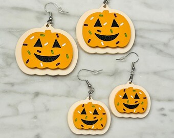 Könnte beinhalten: Drei Paar Halloween-kürbisohrringe. Jedes Paar zeigt einen weißen Kürbis mit einem lächelnden Gesicht und orangefarbenen und grünen Streuseln. Die Kürbisse sind aus Holz und haben einen cremefarbenen Hintergrund.