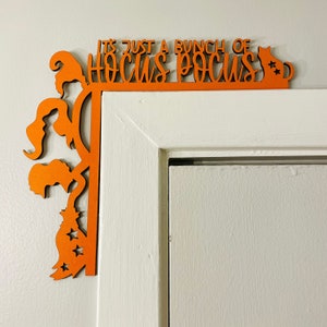 Op de afbeelding: Oranje houten Halloween-deurframe-decoratie. De zin "IT'S JUST A BUNCH OF HOCUS POCUS" is uit het hout gesneden. De decoratie toont silhouetten van heksen, een kat en sterren.
