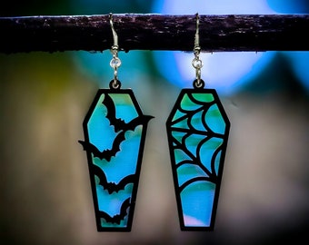 May include: A pair of black coffin-shaped earrings with a blue and green iridescent finish. One earring features a bat design, while the other features a spiderweb design.