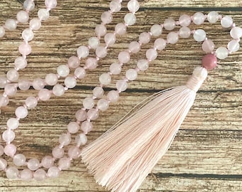 May include: A pink rose quartz mala bead necklace with a tassel. The necklace features round, light pink beads strung together with pink thread. A larger pink bead and a tassel are at the end of the necklace. The background is a wooden surface.