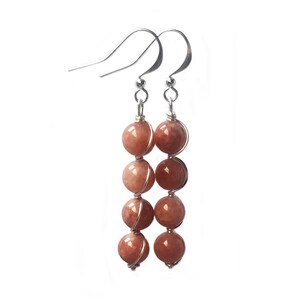 May include: A pair of silver dangle earrings with five peach-colored gemstone beads on each earring. The beads are wrapped in silver wire.
