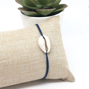 Peut inclure: Un bracelet élastique bleu marine avec un charme en coquillage de cowrie blanc. Le bracelet est posé sur une surface en tissu beige.