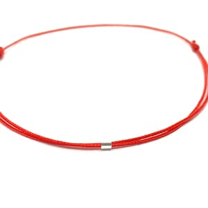 Peut inclure: Un bracelet minimaliste en cordon rouge avec une petite perle cylindrique de couleur argentée. Le bracelet est noué aux deux extrémités et est posé sur un fond blanc. Ce design simple convient au quotidien.