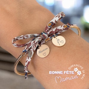 Peut inclure: Bracelet jonc doré avec un nœud en ruban à motifs floraux. Deux breloques rondes dorées sont attachées, l'une avec le mot "Maman" et un cœur, l'autre avec "Je T♡". Le texte "BONNE FÊTE Maman" est également visible.
