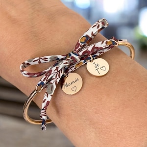 Peut inclure: Bracelet jonc doré orné d'un nœud en tissu fleuri. Deux breloques rondes dorées sont attachées, l'une avec le mot "Mamie" et un cœur, l'autre avec "Je T♡". Le nœud en tissu présente un motif de fleurs rouges, bleues et jaunes sur fond blanc.