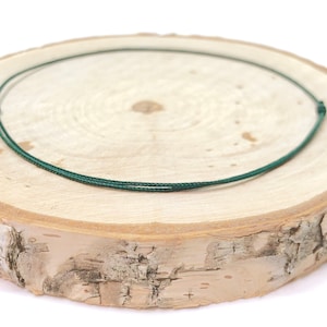 Peut inclure: Un collier vert foncé au design simple, posé sur une surface en bois brun clair. Le collier est fait d'un matériau fin semblable à un cordon et est noué aux extrémités. La surface en bois a un aspect naturel et organique.