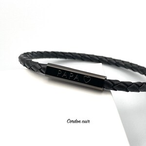 May include: Black leather bracelet with a black metal bar engraved with "PAPA" and a heart symbol. The bracelet is braided and has a simple design.