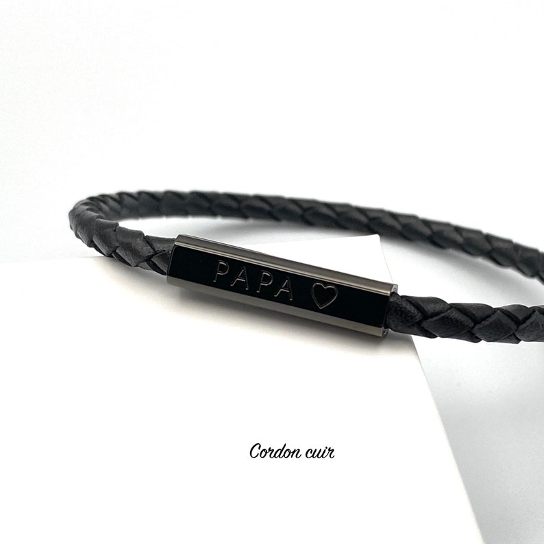 May include: Black leather bracelet with a black metal bar engraved with "PAPA" and a heart symbol. The bracelet is braided and has a simple design.